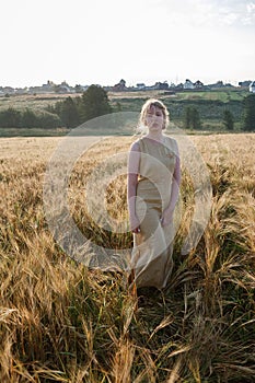 Young pretty girl in yellow dress stands at field of ears in rays of rising sun. grove and village in background