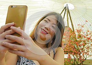 Young pretty and happy Asian Korean tourist woman smiling holding mobile phone taking self portrait selfie picture outdoors in bea