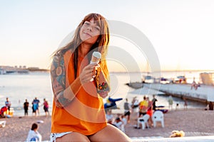 Young pretty happy Caucasian woman with tattoos holds ice cream at sunset beach. Concept of freedom and summer vacation