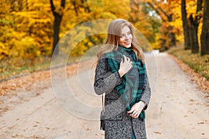 Young pretty happy smiling european woman in an elegant gray coat with a fashionable green warm scarf walks in a golden park