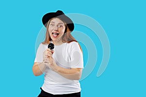 Young pretty happy woman in black hat singing a song with a microphone, isolated on blue background, studio portrait.