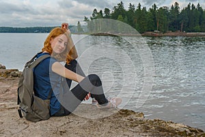 Young pretty lady with backpack sitting on bay shore in summer evening. Tourist on the beautiful landscape background. Monrepos