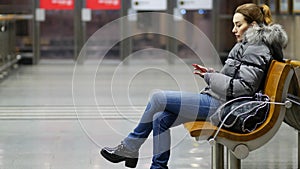 Young pretty lady sits on wooden bench in spacious lit hall with smartphone