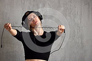 A young pretty long-haired DJ girl in a black top, black baseball cap and black headphones stands against a gray wall