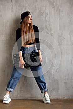 A young pretty long-haired DJ girl in a black top, blue loose jeans and a black baseball cap stands against a gray wall