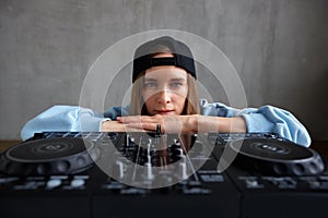 A young pretty long-haired DJ girl in a blue sweater and black baseball cap poses with a black DJ mixing console and