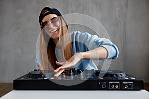 A young pretty long-haired DJ girl in a blue sweater, black baseball cap and sunglasses poses and mixes music tracks on