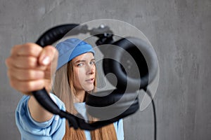 A young pretty long-haired DJ girl in a blue sweater and a funny blue hat holds black headphones in outstretched hands