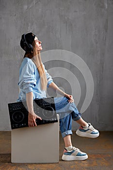 A young pretty long-haired DJ girl in a blue sweater, jeans, a black baseball cap and black headphones sits with a black