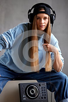 A young pretty long-haired DJ girl in a blue sweater, jeans, a black baseball cap and black headphones sits with a DJ