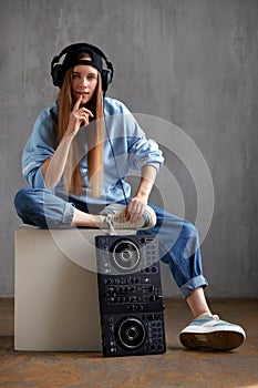 A young pretty long-haired DJ girl in a blue sweater, jeans, a black baseball cap and black headphones sits with a DJ