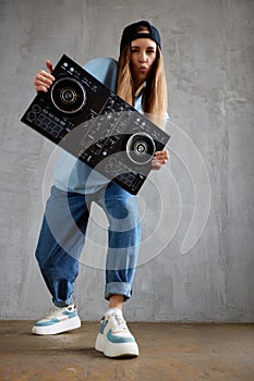 A young pretty long haired DJ girl in a blue sweater, jeans and a black baseball cap poses with a black DJ mixing