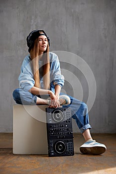 A young pretty long haired DJ girl in a blue sweater, jeans and a black baseball cap is posing while sitting on a white