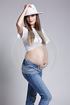 Young pretty pregnant woman in white t-shirt, hat and jeans. Female with hand on belly exposed. 5th month of pregnancy