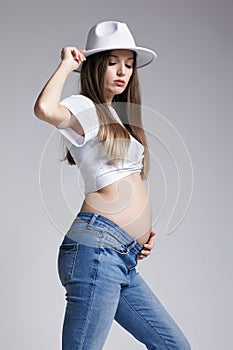 Young pretty pregnant woman in white t-shirt, hat and jeans. Female looking at pregnant belly. Belly exposed. 5th month of