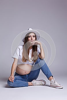 Young pretty pregnant woman in white t-shirt, hat and jeans sit on the floor on background. Female with belly exposed. 5th month