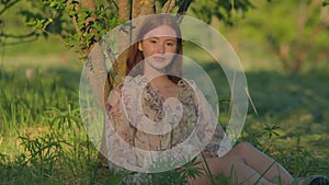 Young pretty redhead girl relax under tree sitting on green meadow in sunshine in park in summertime. Natural beauty