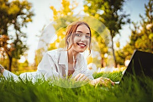 Young pretty redhead positive girl uses laptop pc and headphones while laying on the grass in park
