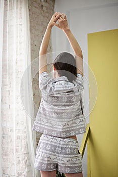 Young pretty slim brunette woman, wearing grey white pajamas pajamas with christmas pattern, with her back to the camera,