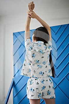Young pretty slim brunette woman, wearing white pajamas,with blue flowers pattern,with her back to camera, stretching in the