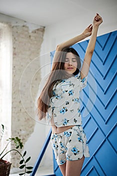 Young pretty slim brunette woman, wearing white pajamas,with blue flowers pattern, stretching in the morning. in light room near