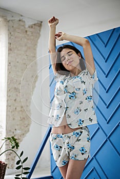 Young pretty slim brunette woman, wearing white pajamas,with blue flowers pattern, stretching in the morning. in light room near