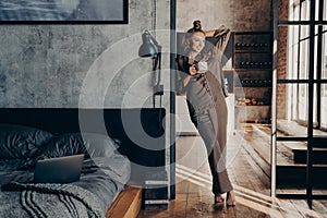 Young pretty slim woman wearing brown satin pajama holding coffee cup while standing in bedroom