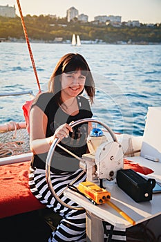 Young pretty smiling woman in black shirt and striped skirt driving luxury sailing boat in sea, sunset