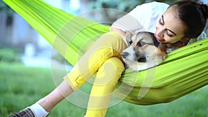 Young pretty smiling woman with cute Welsh Corgi dog lying in bright green hammock in park
