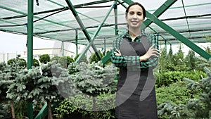 Young pretty smiling woman farm worker standing with crossed hands in greenhouse, looking at camera.