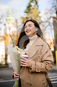 Young pretty smiling woman with long hair, holding a bouquet of lilies and a disposable cup of coffee