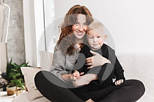 Young pretty smiling woman sitting on sofa with her little handsome son happily looking in camera together. Mom with