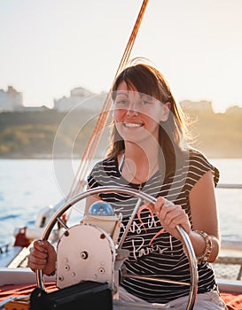 Young pretty smiling woman in striped shirt driving luxury yacht in sea, sunset