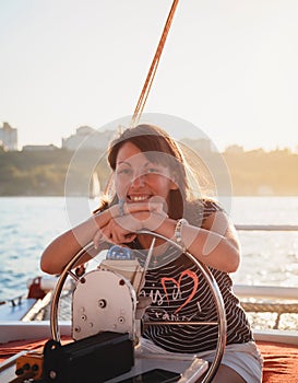 Young pretty smiling woman in striped shirt and white shorts driving luxury yacht in sea, hot summer day, sunset