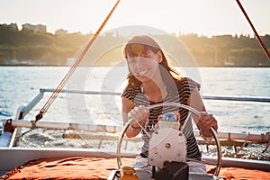 Young pretty smiling woman in striped shirt and white shorts driving luxury yacht in sea, sunset