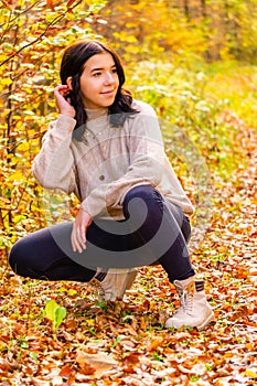 Young pretty stylish dressed girl with beige sweater and black jeans sits in the colorful autumn forest in October and brushes her