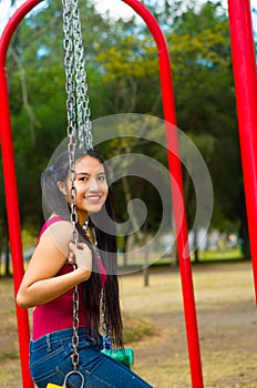 Young pretty teenage girl with pig tails wearing jeans and purple top, sitting on swing at outdoors playground, smiling