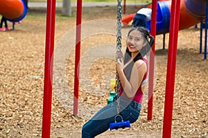 Young pretty teenage girl with pig tails wearing jeans and purple top, sitting on swing at outdoors playground, smiling