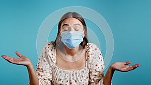 Young pretty unsure woman in protective mask shrugs arms, makes gesture of I don`t know, can`t help anything. Blue
