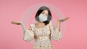 Young pretty unsure woman in protective mask shrugs arms, makes gesture of I don`t know, can`t help anything. Pink