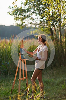 Young pretty woman artist draws paints a picture of lake on open plain air outdoors