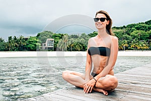 Young pretty woman in black bikini sitting alone on the pier near the sea