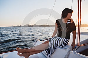 Young pretty woman in black shirt and striped skirt at luxury yacht in sea, sunset