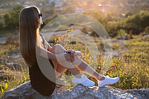 Young pretty woman in black short summer dress sitting on a rock relaxing outdoors at sunset. Fashionable female enjoying warm