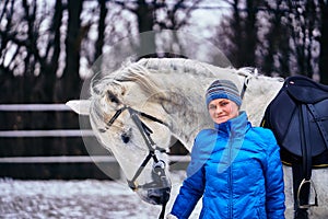 Young pretty woman in blue jacket and sports hat on a walk with a white horse on a winter cloudy day.