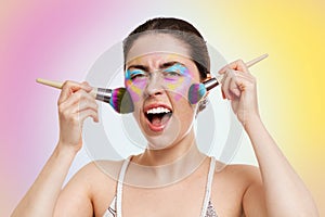 A young pretty woman with bright eyelid makeup, holds makeup brushes near her cheeks and screams. Yellow-pink background. Copy