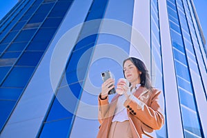 Young, pretty woman in brown coat and glass of coffee in hand, checking social networks on her cell phone on her way out of the