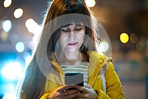 Young pretty woman browsing internet on her mobile phone on a city street at night outdoors