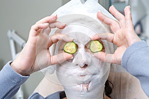 A young pretty woman with a cream mask on her face holds a plastic cucumber in her hands