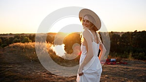 Young pretty woman in dress and hat outdoors in field with amazing sunset view enjoying a warm summer day.
